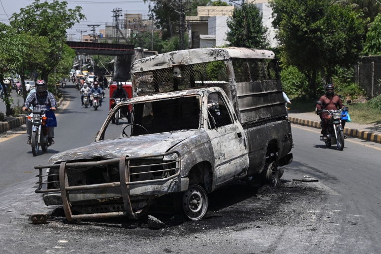 Commuters walk past a burnt and torched vehicle during a protest by Pakistani Tehreek-e-Insaf party activists and supporters of former Pakistani Prime Minister Imran Khan over the arrest of their leader